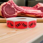 Tri-Tip Butcher sticker red identification label with black print on a roll of stickers with meat in background